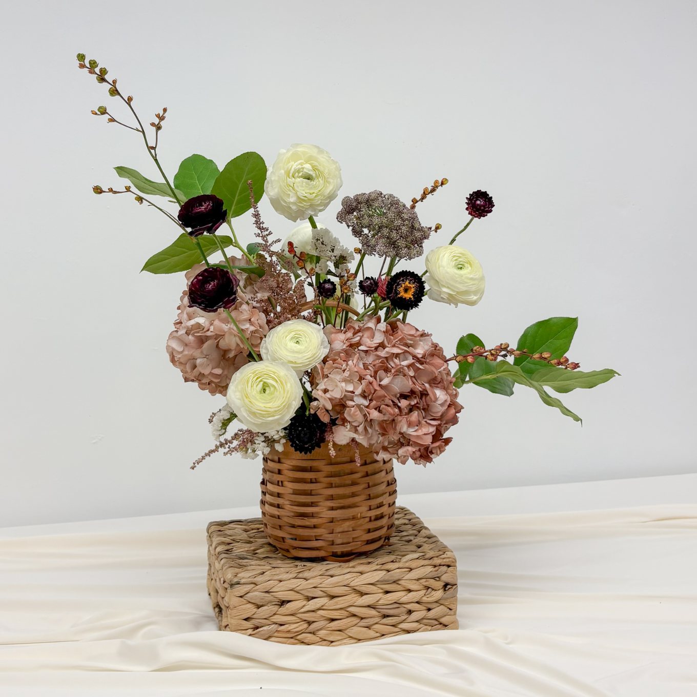 Ivory ranunculus, dusty pink hydrangea, burgundy ranunculus, and strawflowers in a woven basket on a woven stand